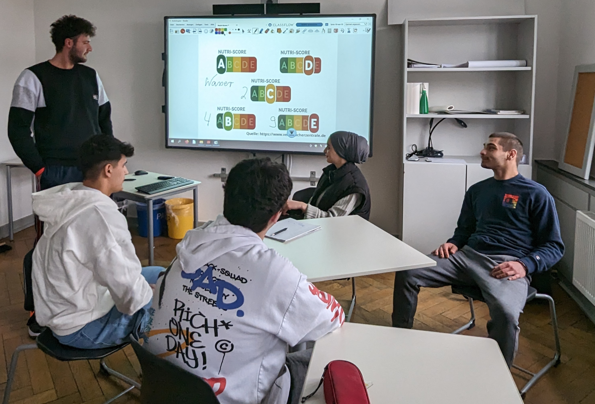 Fünf junge erwachsene sitzen zusammen vor einem Smartboard. Darauf sind Abbildungen des Nutri-Scores zu sehen. Einer der jungen Erwachsenen steht neben der Tafel und spricht zu den anderen.