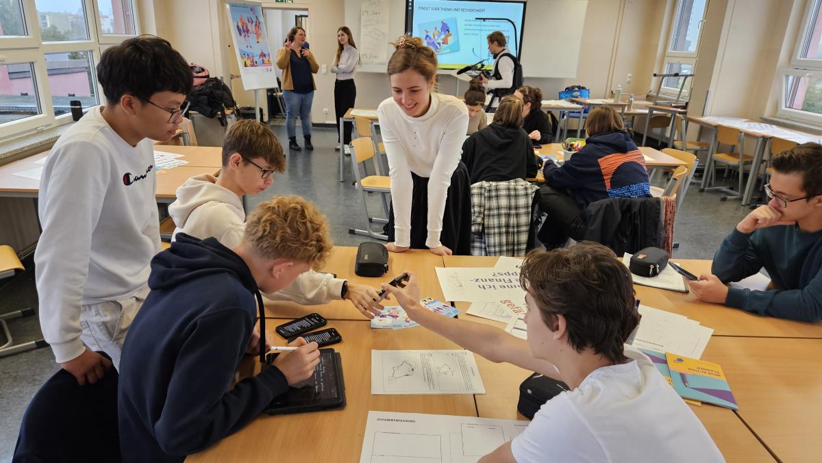 Junge Leute in einem Klassenraum arbeiten konzentriert an Projekten. Unterstützt werden sie von Trainerinnen und der Klassenlehrerin. Ein Kameramann filmt im Hintergrund.